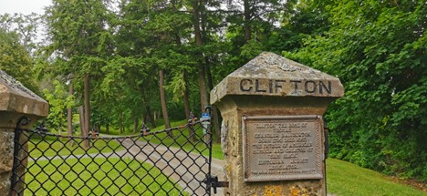 Photo of Clifton Park in mid-summer with overcast sun. Two stone gate posts stand with a black chain link fence between them. Clifton is carved in thick letters along the top of one.  Underneath a plaque with small, unreadable lettering.  Behind the gate a driveway stretches into the background, with large deep green hemlock trees and rolling green hills.  