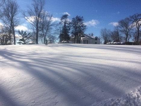Photo of Clifton Museum Park in winter on a sunny day with blue sky. In the foreground the lawn is covered in smooth white snow and long shadows from nearby bare trees. In the background the large, old, white house is covered in snow.