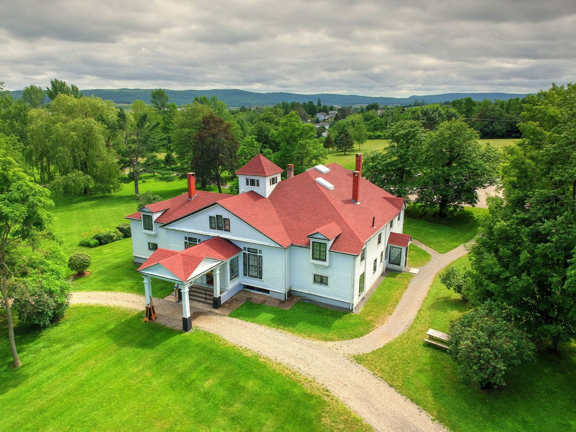 An aerial view of Clifton House, a white building with a red roof, surrounded by green lawns, trees, and walking paths under a cloudy sky.