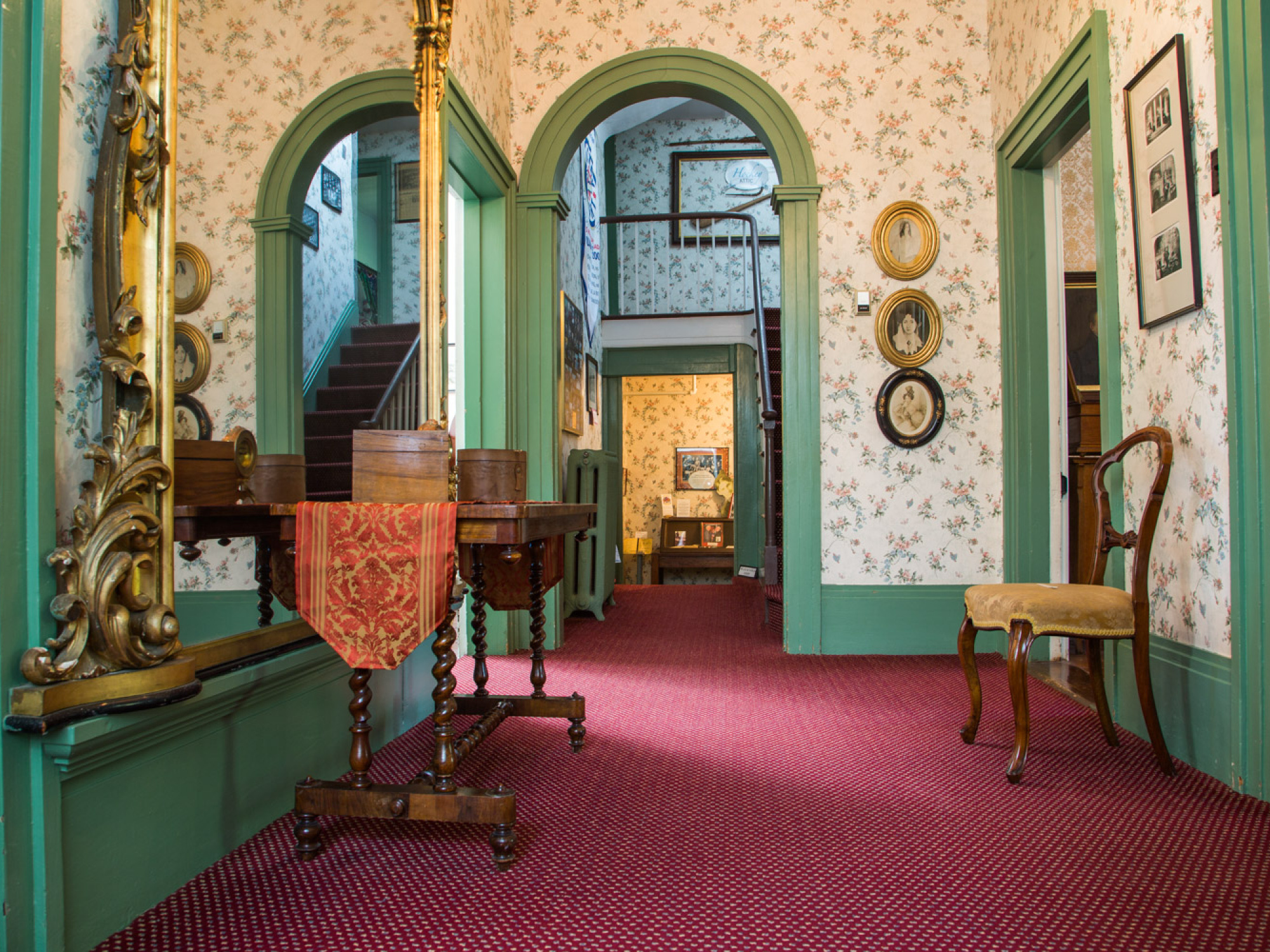 Interior shot of a hallway with green-painted walls, a red patterned carpet, antique wooden furniture, and framed artwork, leading to a staircase in the distance.