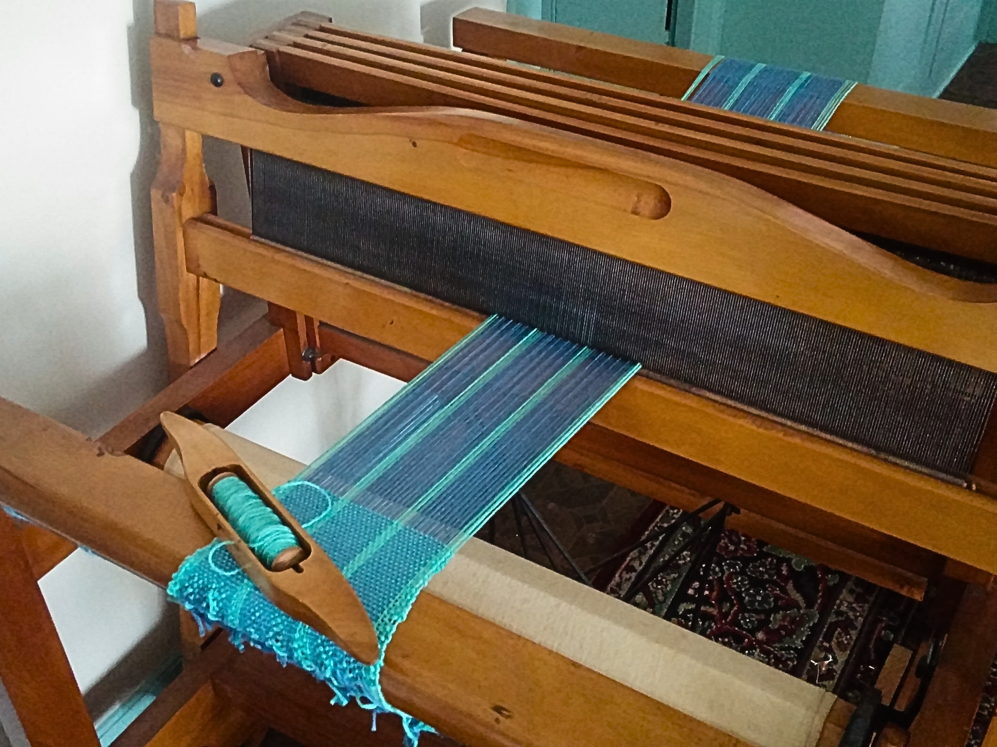 A wooden loom with a partially woven piece of fabric featuring blue and turquoise threads, and a shuttle with yarn visible.