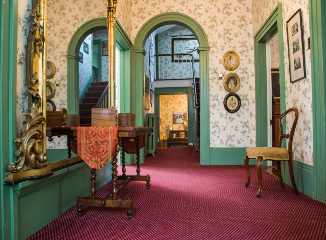 Interior shot of a hallway with green-painted walls, a red patterned carpet, antique wooden furniture, and framed artwork, leading to a staircase in the distance.