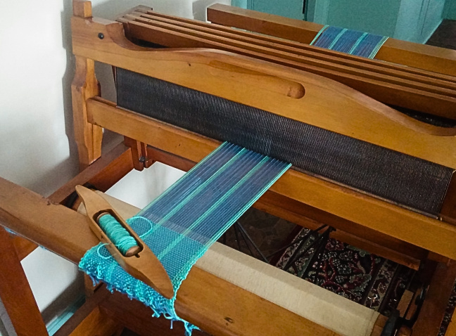 A wooden loom with a partially woven piece of fabric featuring blue and turquoise threads, and a shuttle with yarn visible.